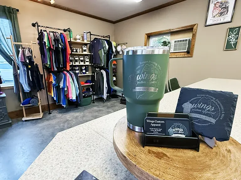 A Wings Custom Apparel cup on a table with a business card and coaster in a room with blank t-shirts.